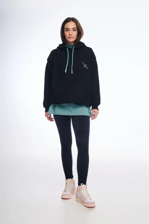 Person wearing a black hoodie with a logo, light green shirt, black leggings, and white sneakers on a white background