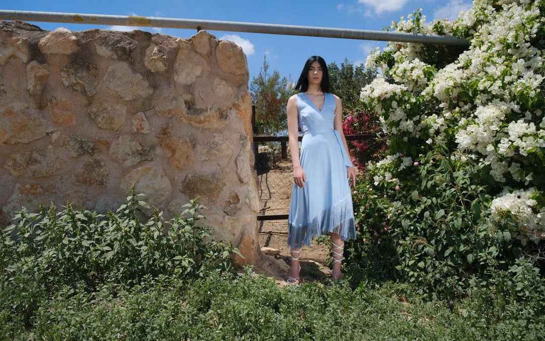 Woman in a light blue dress standing in a garden with stone wall and white flowers.
