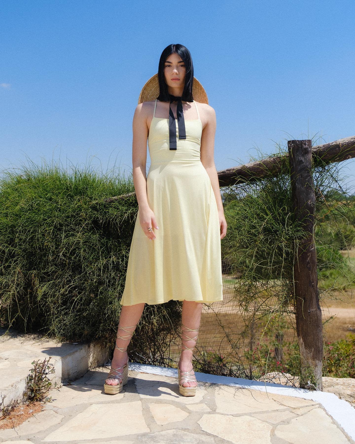 Woman in a yellow dress standing outdoors with a clear blue sky and greenery.