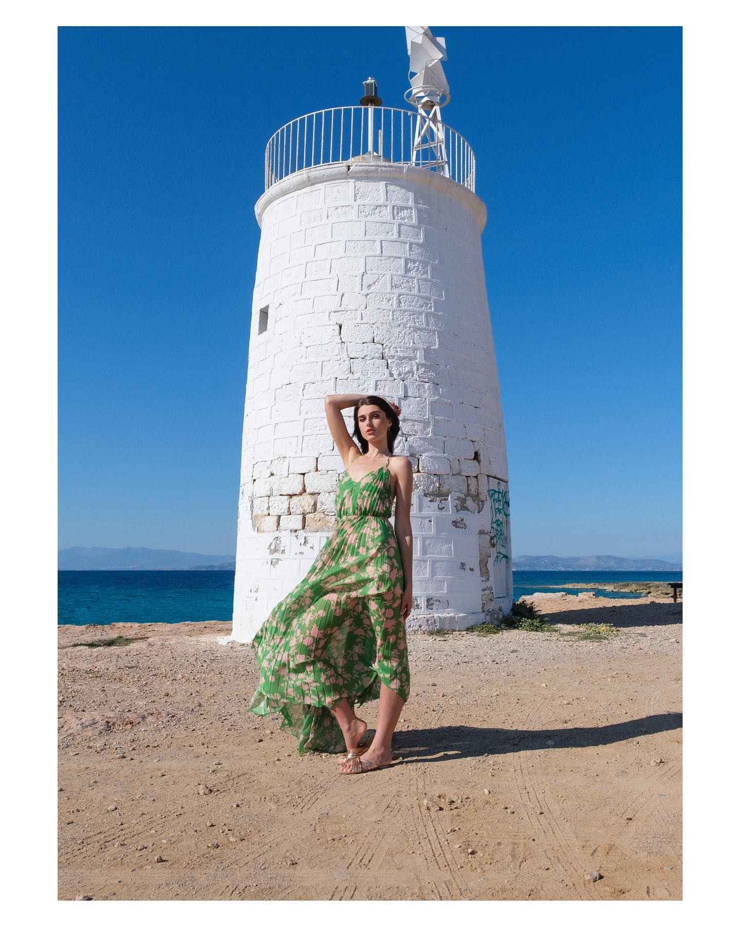 Woman in a green dress standing in front of a white lighthouse by the sea.