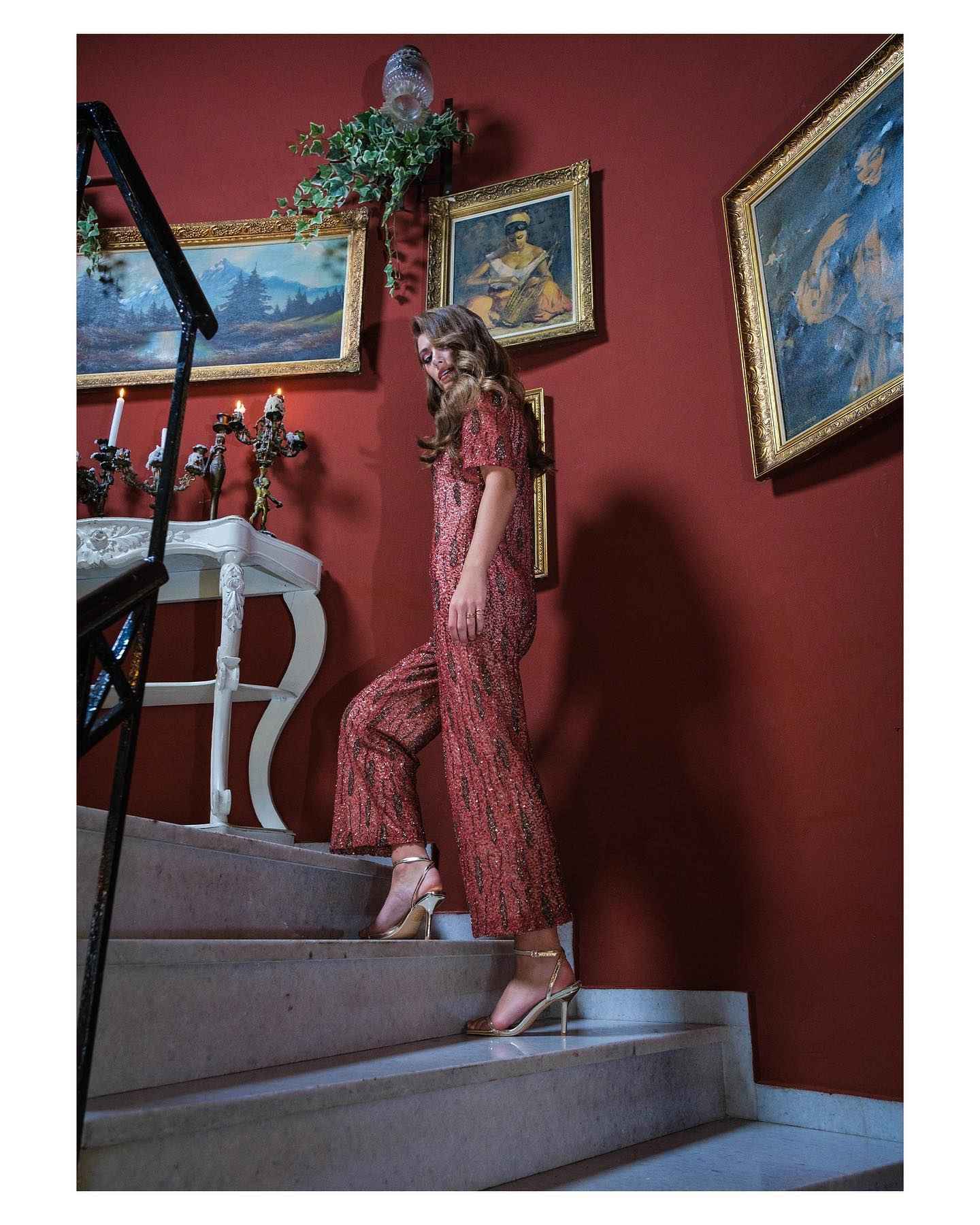 Woman in a red patterned dress standing on a staircase with red walls and framed paintings.