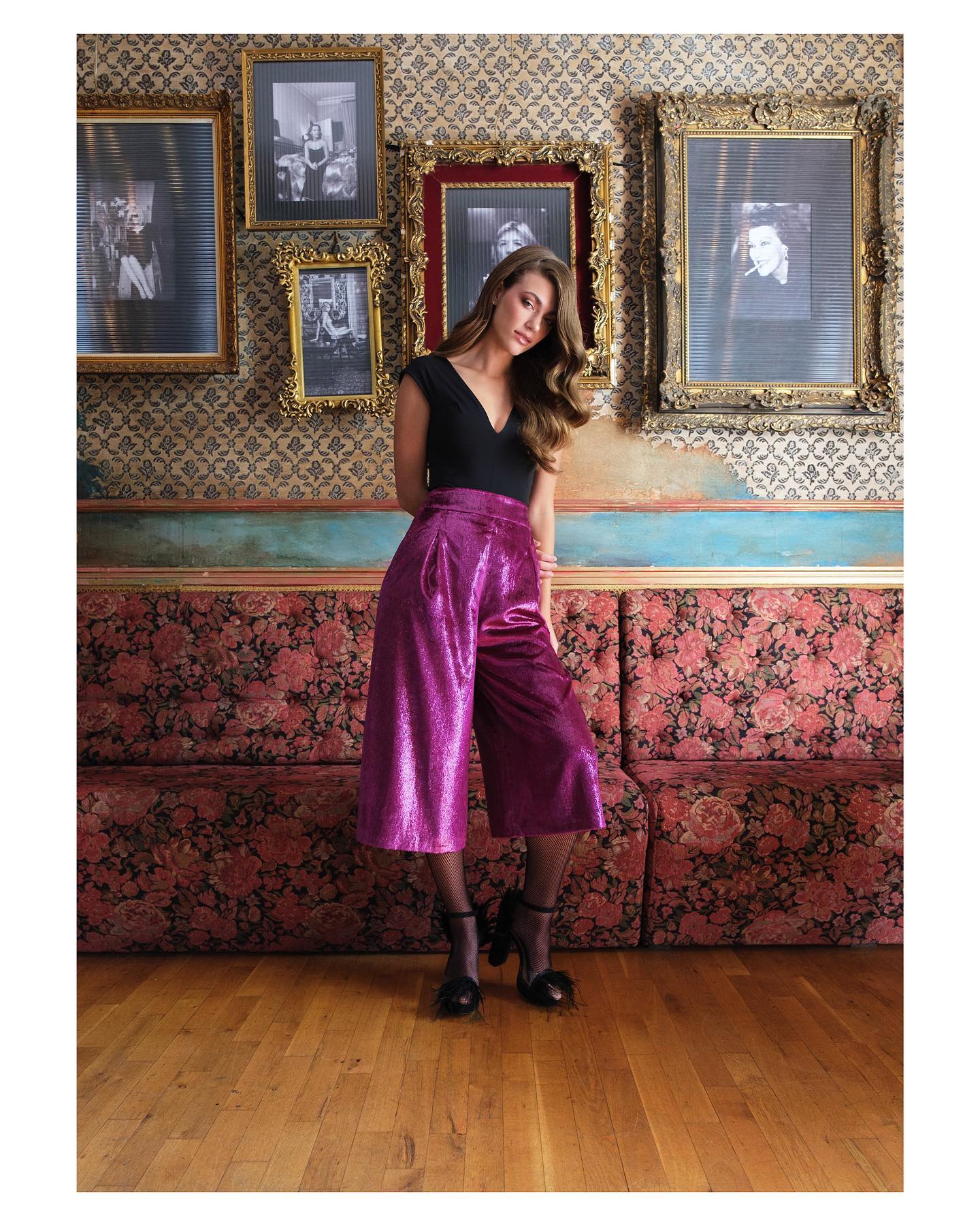 Woman in a black top and pink pants standing in a room with floral wallpaper and framed pictures.