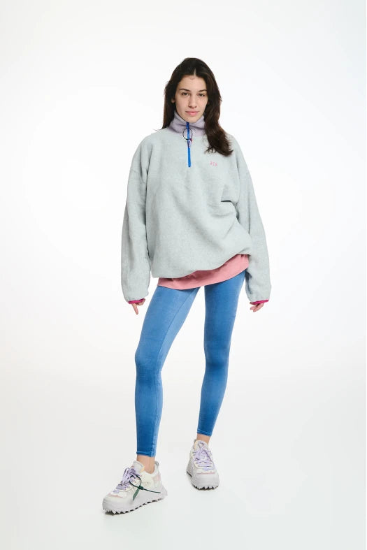 Person wearing a light gray oversized sweater with a pink shirt underneath, blue leggings, and white sneakers on a white background.