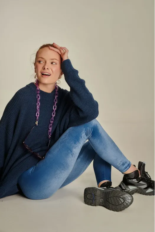Woman sitting on the floor wearing a blue sweater, jeans, and black shoes with a neutral background
