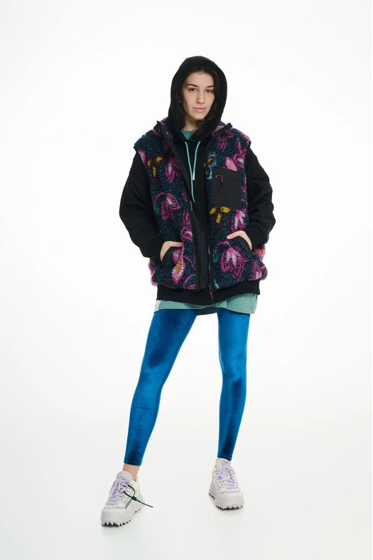 Person wearing a colorful floral-patterned vest over a black hoodie with blue leggings on a white background