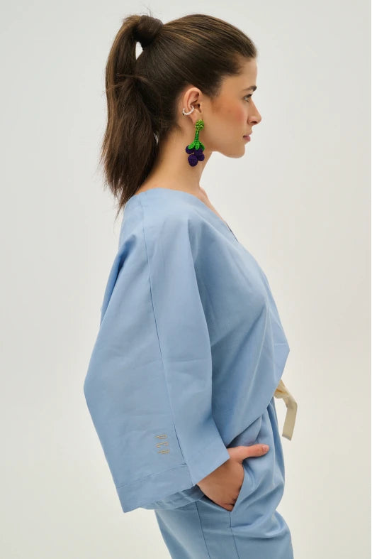Woman wearing a light blue outfit with green and purple earrings on a plain background
