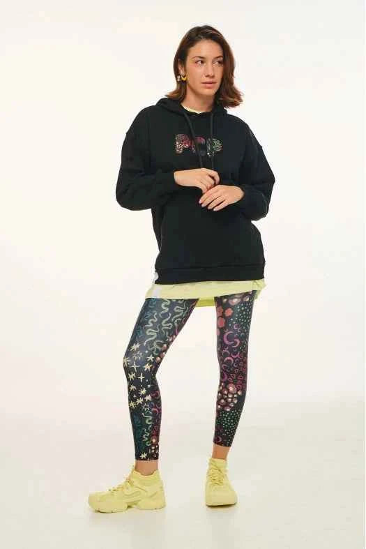 Person wearing a black hoodie with colorful designs, a yellow shirt, patterned leggings, and yellow shoes on a white background.