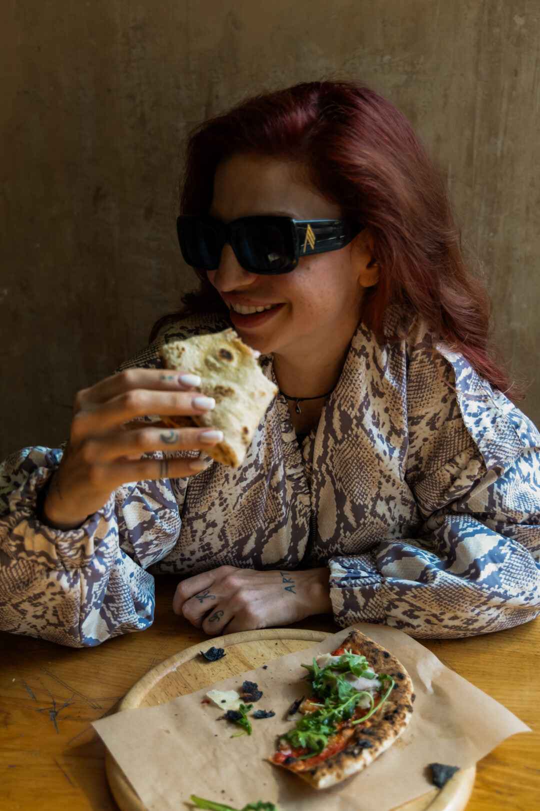 A woman with red hair and sunglasses eating pizza at a wooden table. She is wearing a snakeskin-patterned shirt with long, puffed sleeves. A slice of pizza sits on a wooden board in front of her.