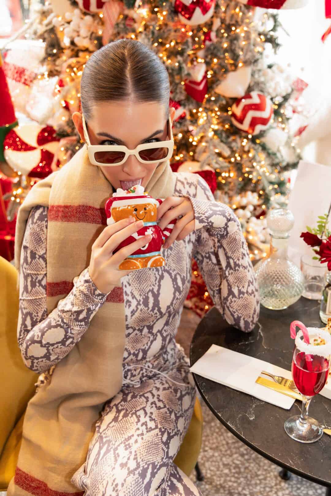 Woman drinking from a festive cup in front of a decorated Christmas tree.