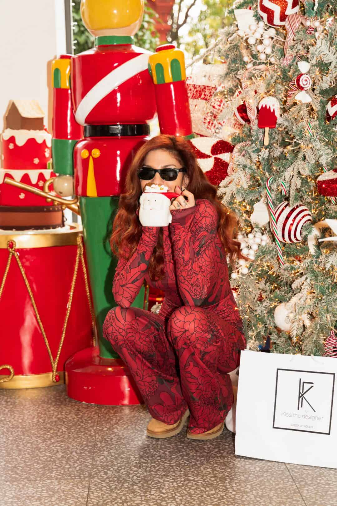 Person in red outfit sitting next to a large nutcracker and Christmas tree, holding a mug.