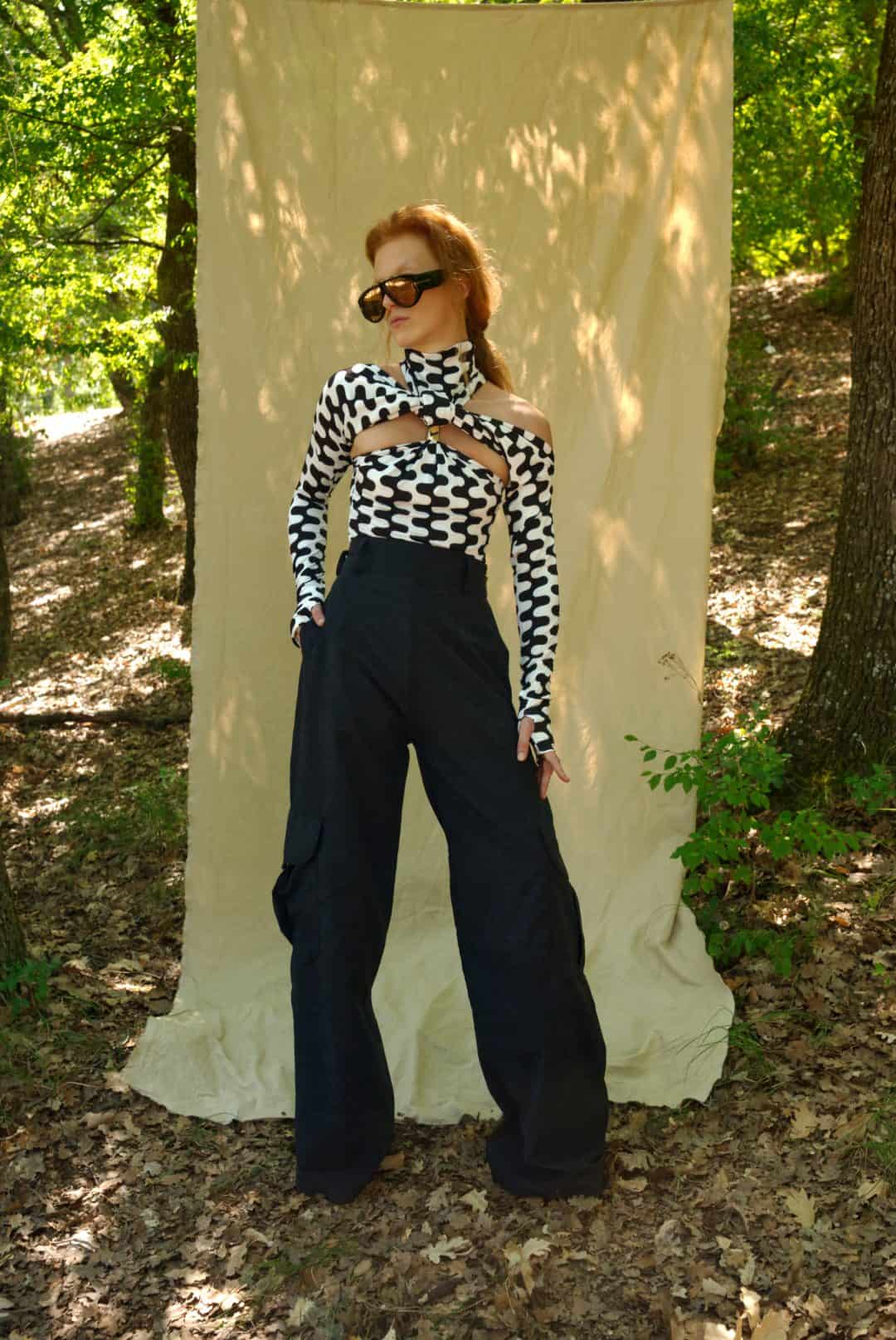 Person wearing a black and white checkered top and black pants standing in front of a white curtain in a forest setting.