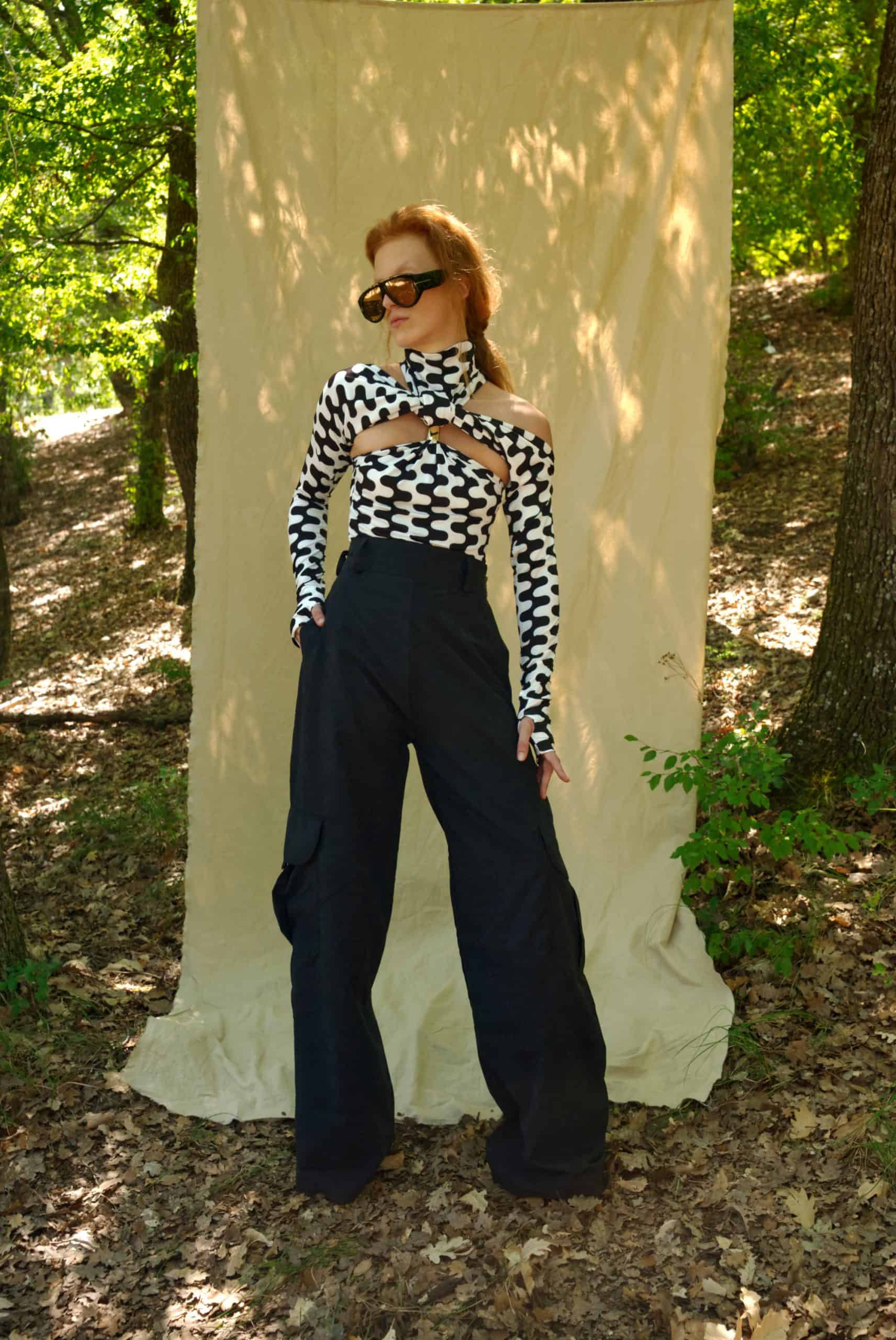 Woman in a black and white checkered top and dark pants standing in front of a white curtain in a forest setting.
