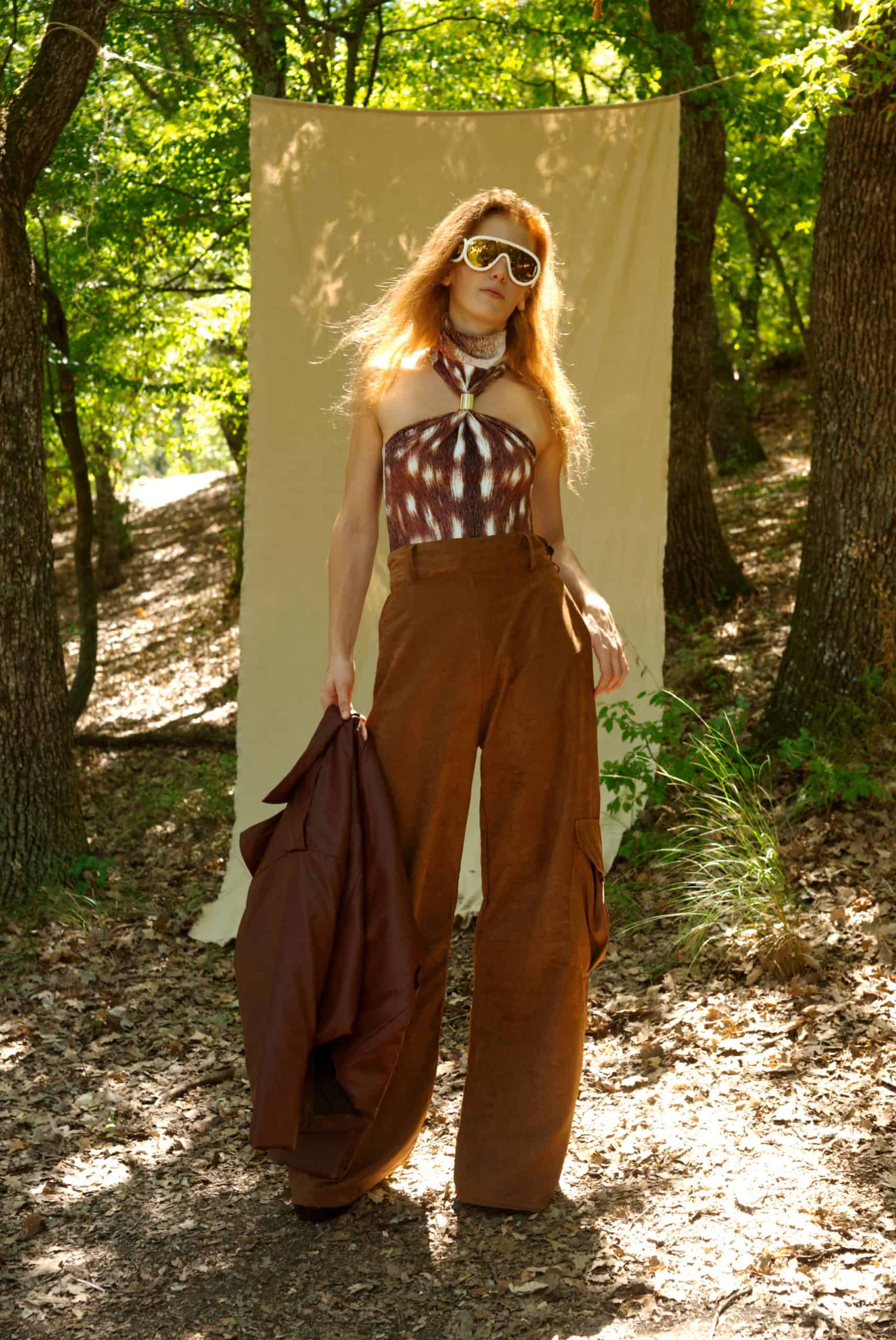 Woman in a patterned top and brown pants standing in a forest with a beige backdrop