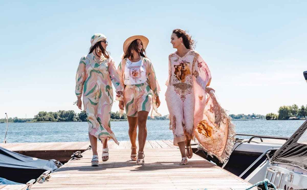 Three women walking on a dock by a body of water, wearing patterned outfits.