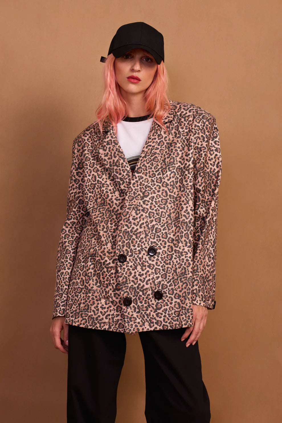 A person wearing a leopard print oversized blazer with black buttons, paired with black pants and a black cap.