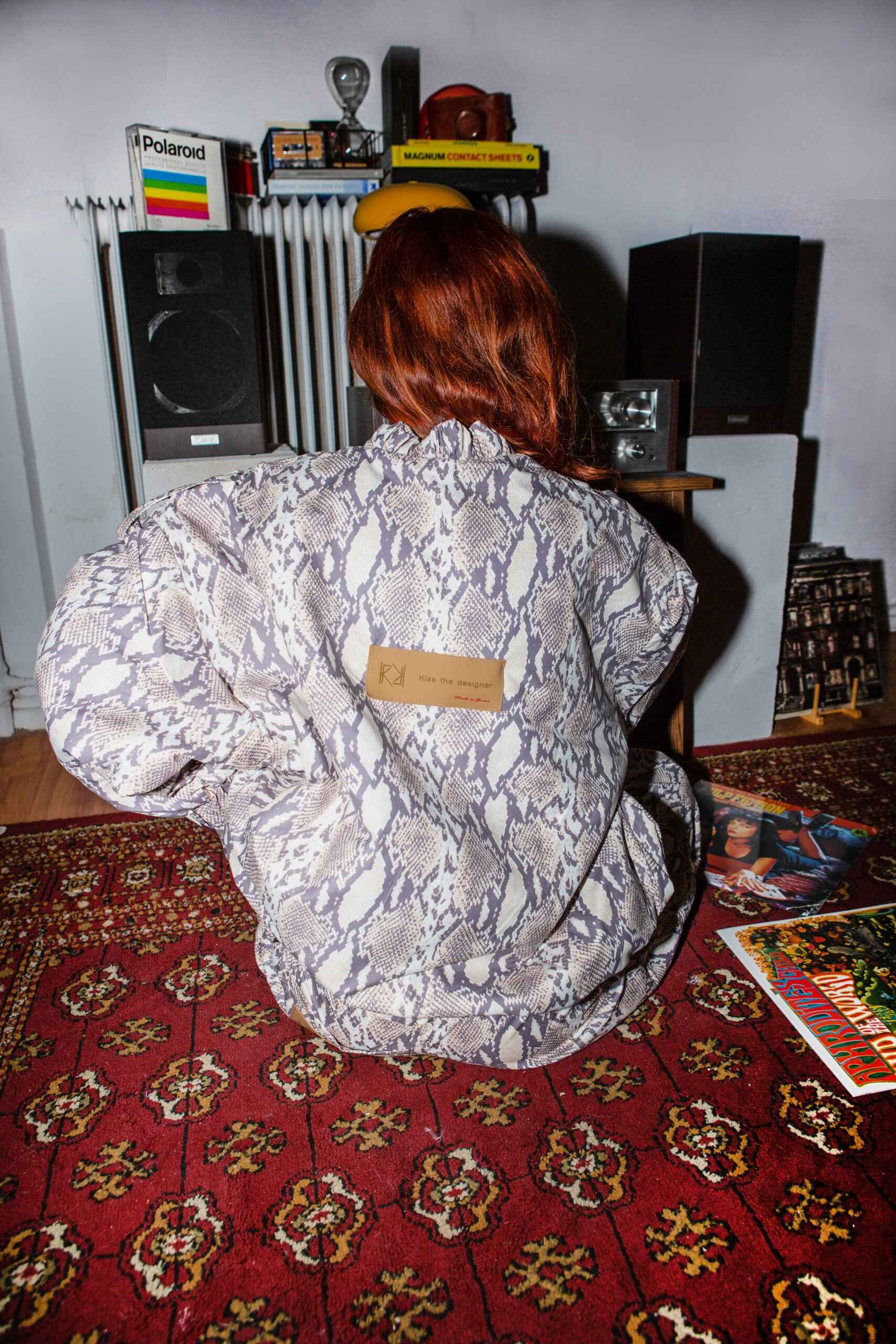 Person sitting on a red patterned rug, wearing a snakeskin-patterned jacket.