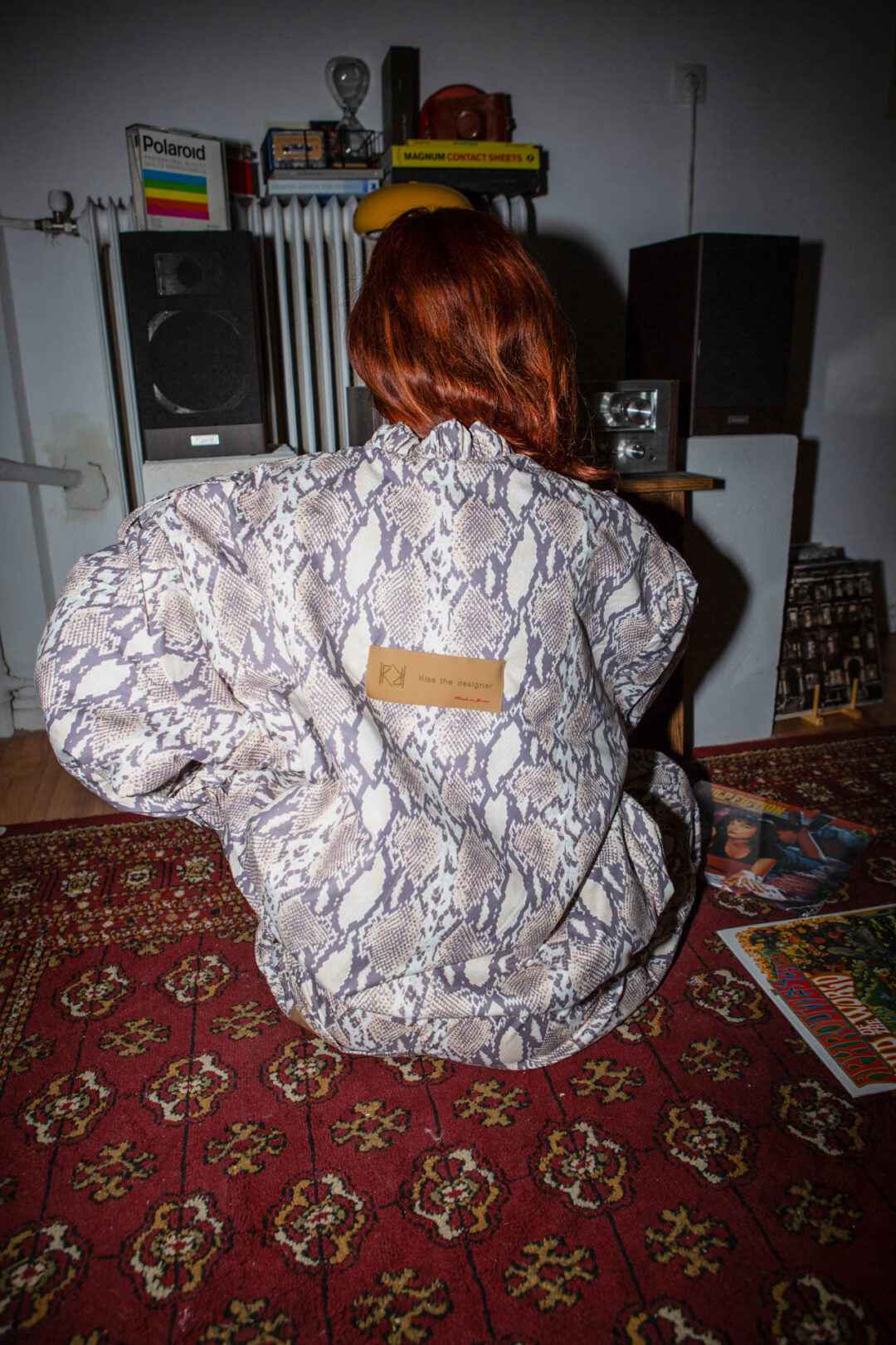 Rear view of person wearing snakeskin-print jacket sitting on red patterned carpet. Speakers, books, and vinyl records in background.