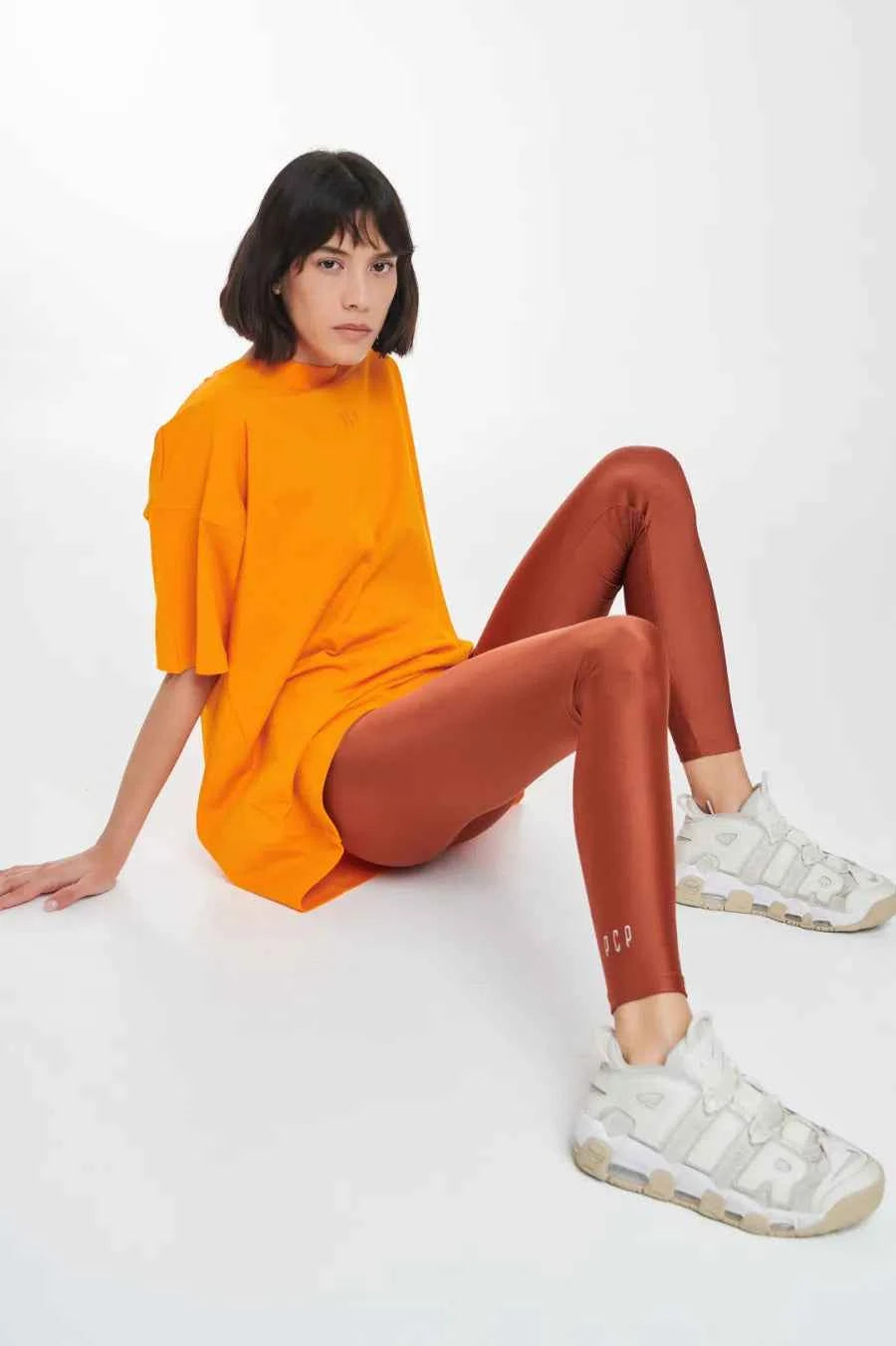 Woman wearing an orange oversized sweater and brown leggings with white sneakers on a white background