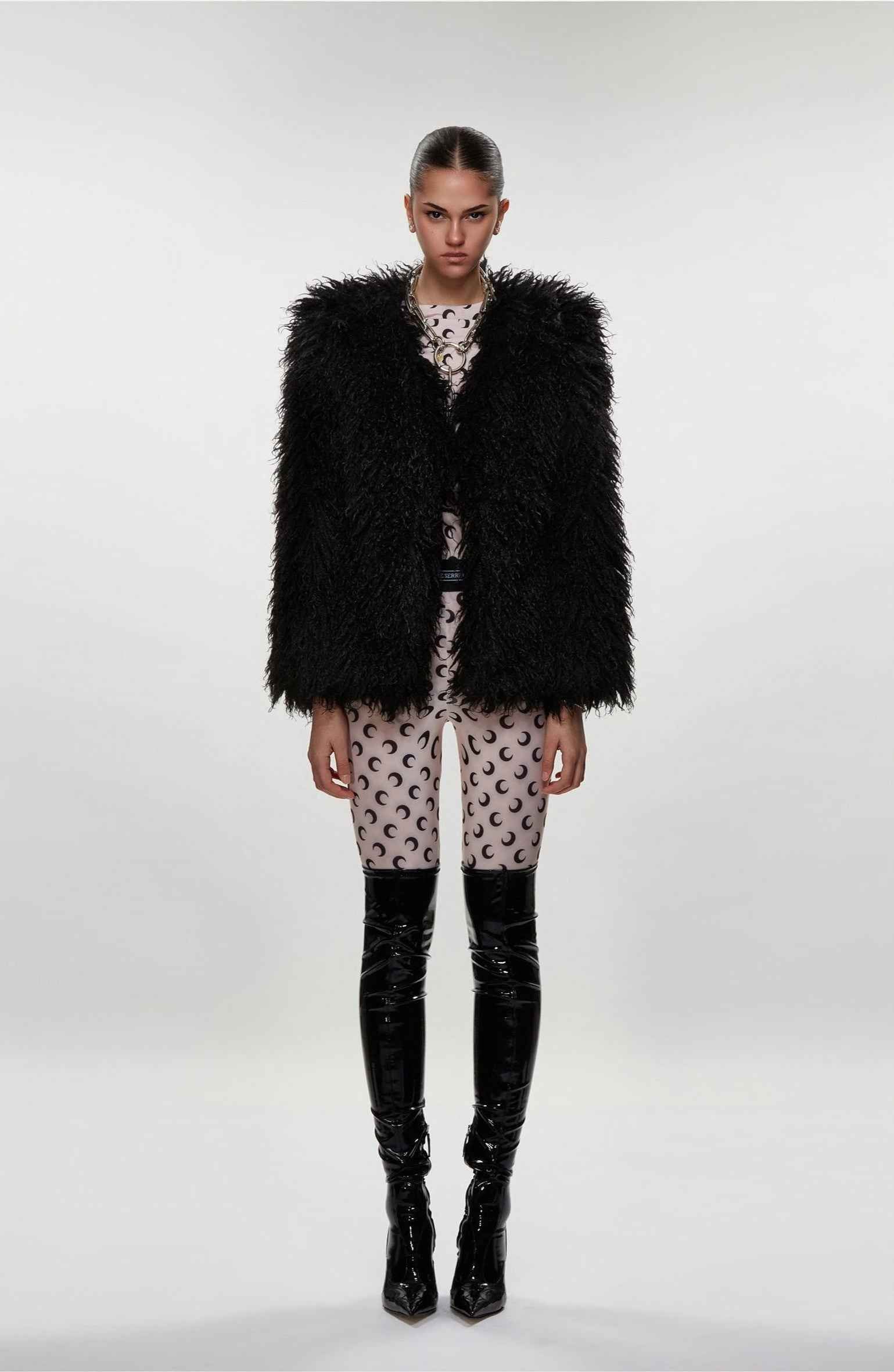 Model wearing a black fur vest, patterned dress, and black knee-high boots on a white background