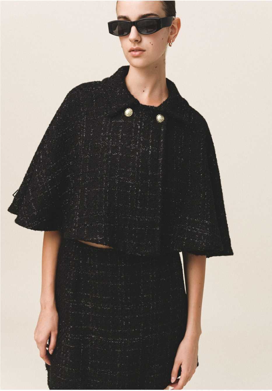 A model wearing a black tweed cape with gold button details and a flared silhouette, paired with a matching skirt.