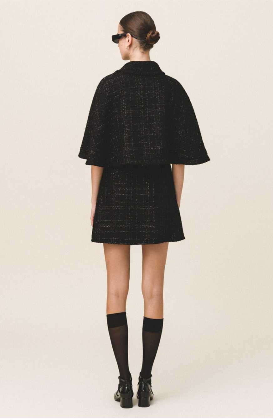 A model wearing a black tweed cape with gold button details and a flared silhouette, paired with a matching skirt.