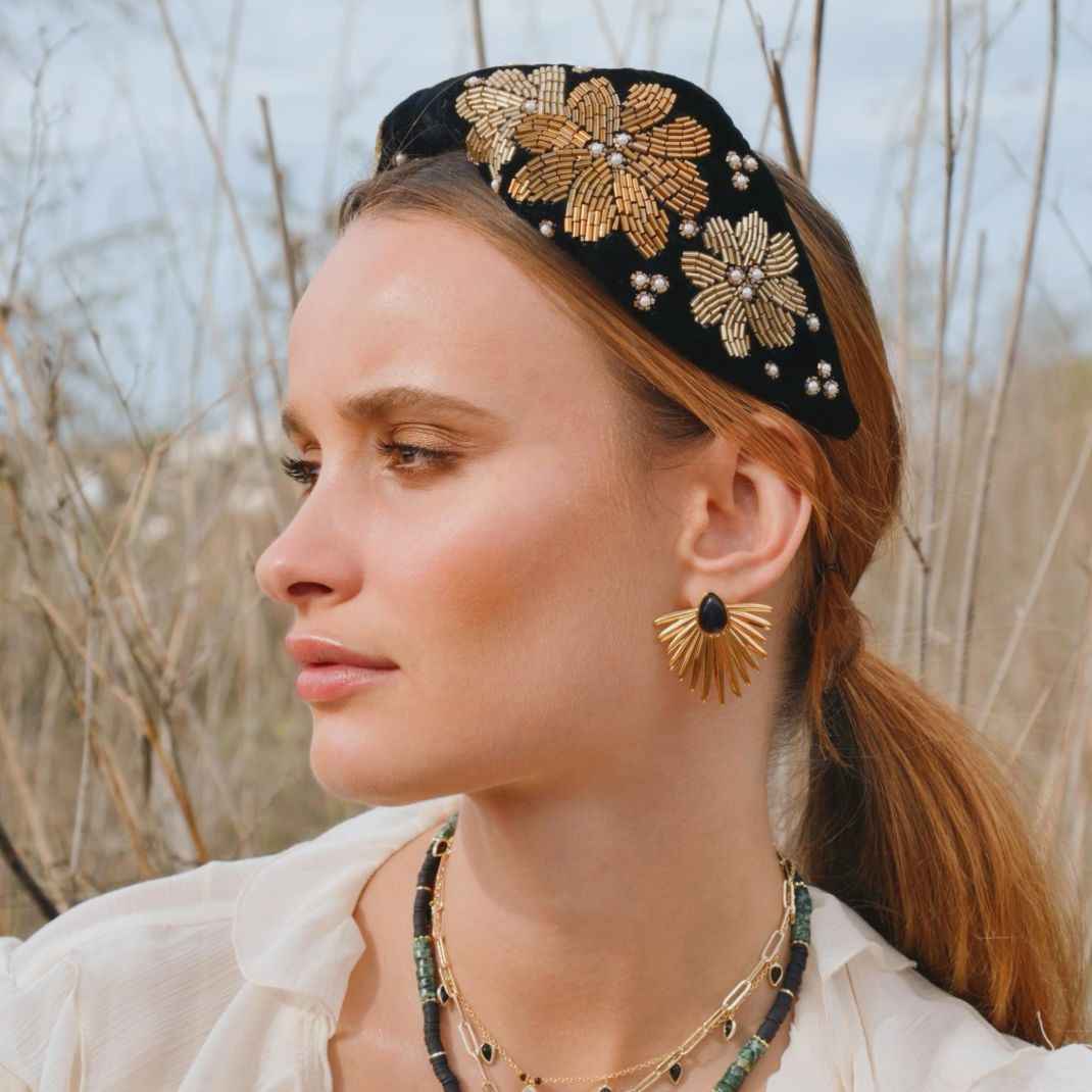 Woman wearing a black headband with gold floral patterns and gold earrings outdoors.