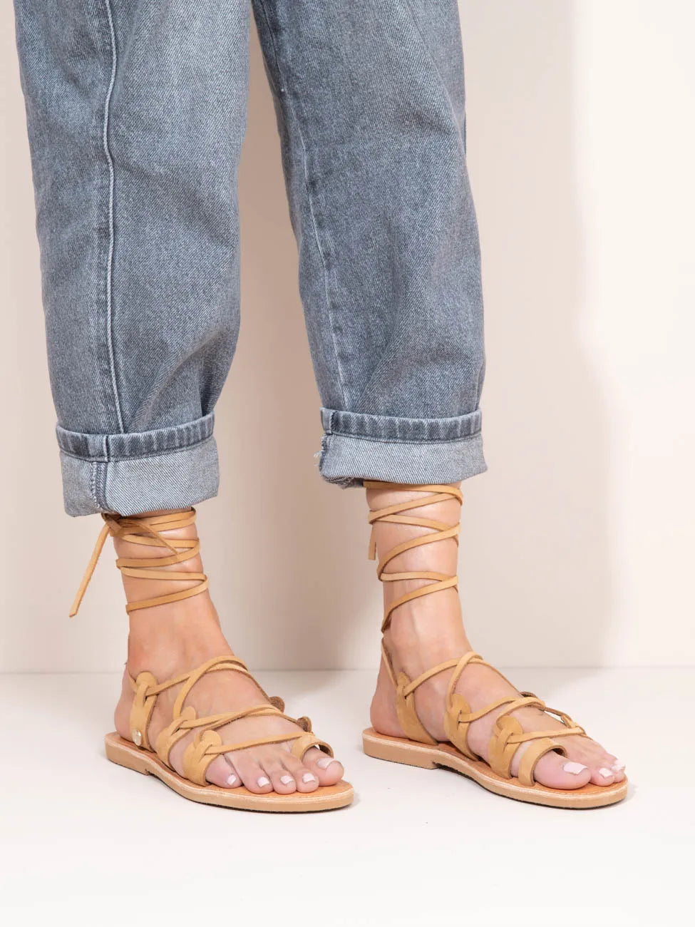 Beige strappy sandals worn with rolled-up blue jeans on a plain background