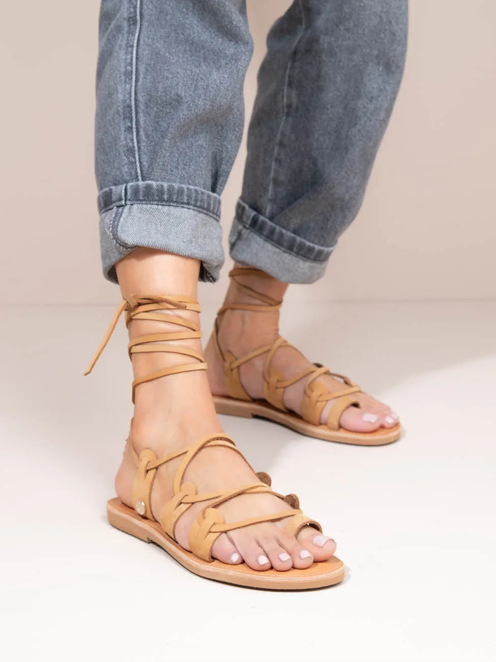 Beige strappy sandals worn with rolled-up blue jeans on a neutral background