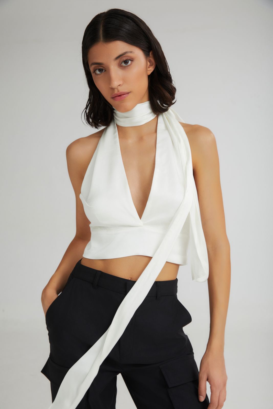 Woman wearing a white halter top and black pants with a white scarf around her neck on a plain background