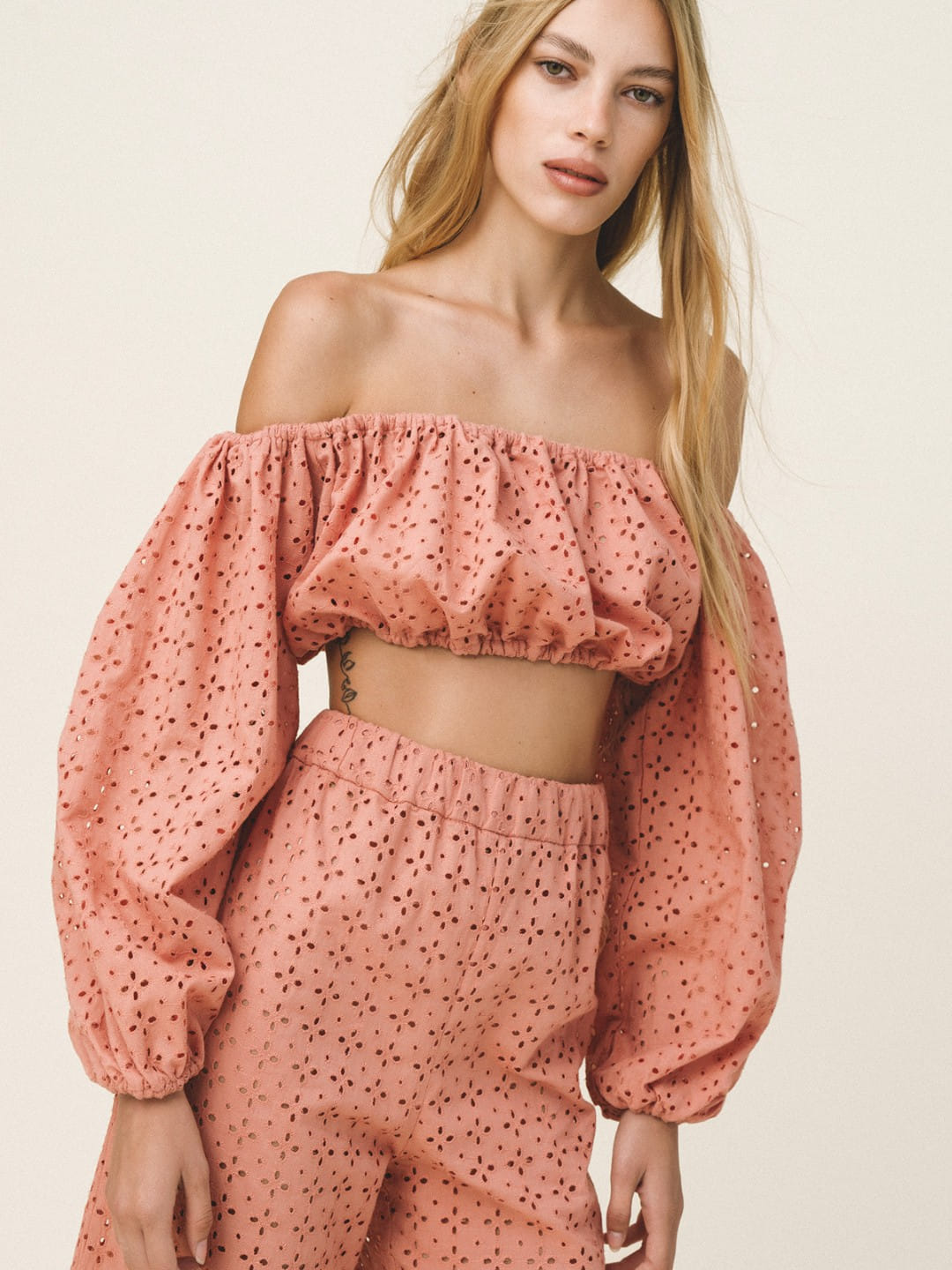 A woman modeling a dusty pink off-the-shoulder crop top and matching high-waisted trousers set, both featuring a small black polka dot pattern, against a solid light beige background.