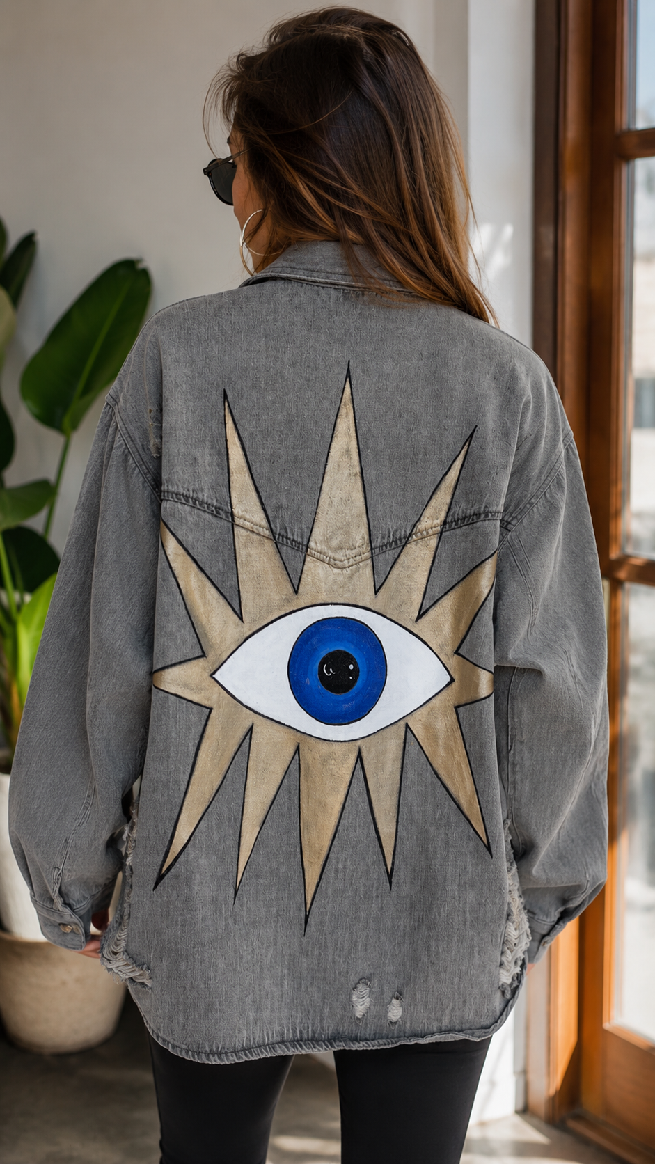 A woman viewed from behind wearing a gray washed denim jacket with a large embroidered or appliqué evil eye and starburst design on the back, paired with black pants in an indoor setting near a window.
