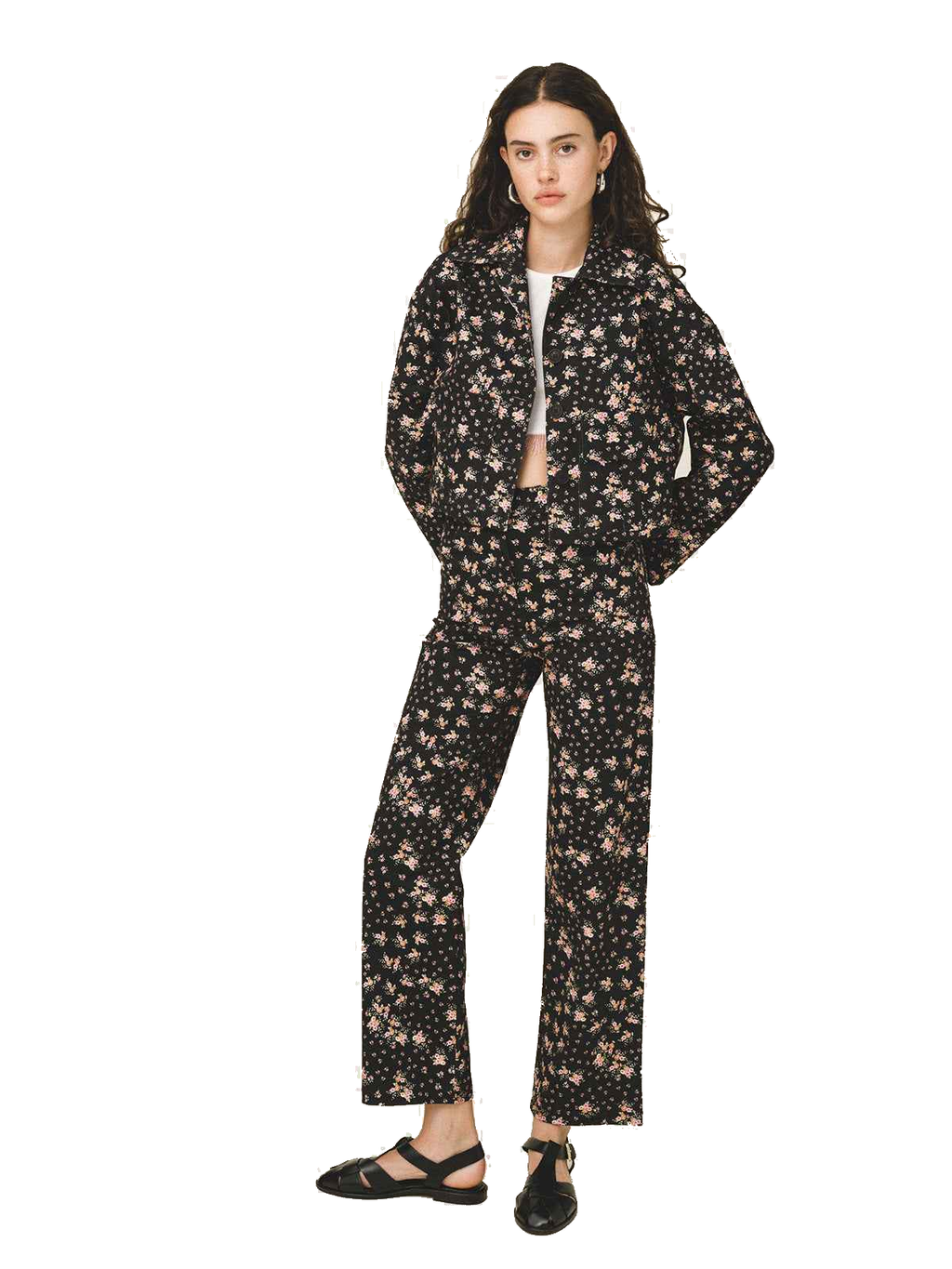 A model stands against a white studio background wearing a matching black two-piece set with a dense beige and pink floral print. The set includes a long-sleeve zip-up jacket and wide-leg trousers, styled with a white undershirt and black flat sandals.