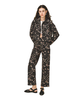 A model stands against a white studio background wearing a matching black two-piece set with a dense beige and pink floral print. The set includes a long-sleeve zip-up jacket and wide-leg trousers, styled with a white undershirt and black flat sandals.