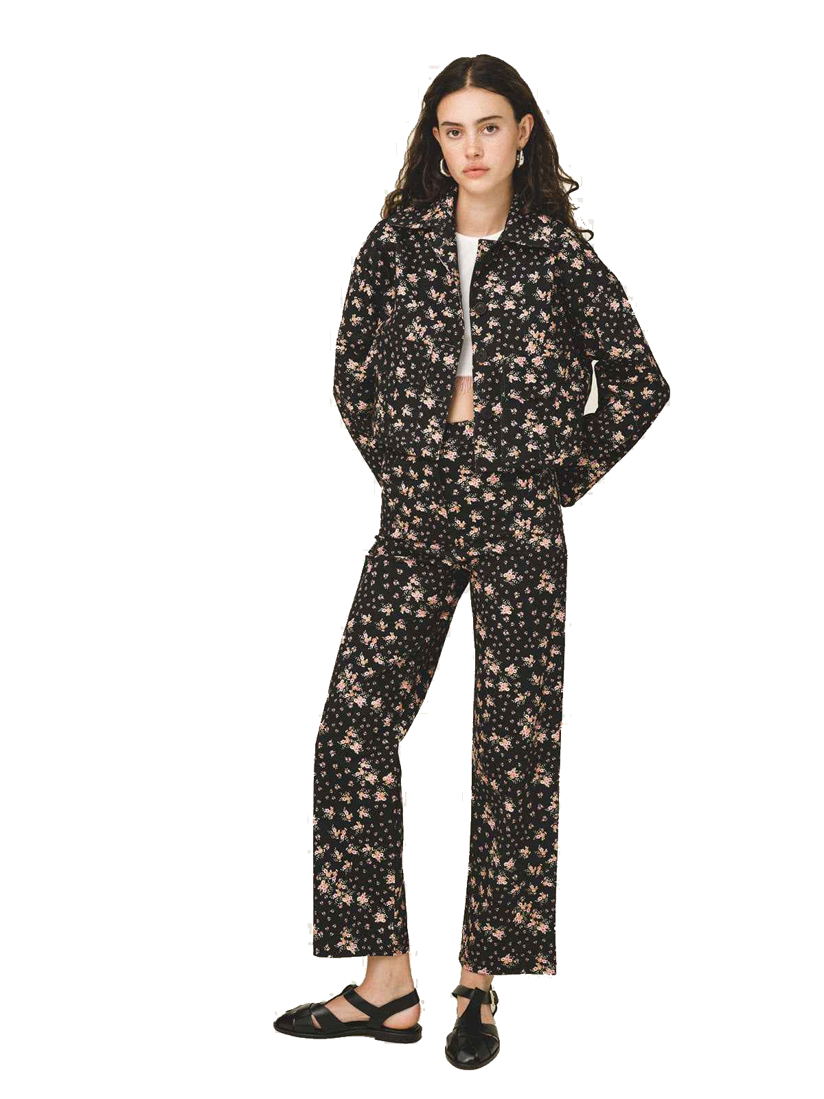A model stands against a white studio background wearing a matching black two-piece set with a dense beige and pink floral print. The set includes a long-sleeve zip-up jacket and wide-leg trousers, styled with a white undershirt and black flat sandals.