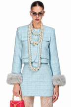 A model wearing a light blue tweed mini skirt suit with faux fur trim, accessorized with multiple layers of pearl necklaces and sunglasses, holding a pink beaded handbag against a white background.