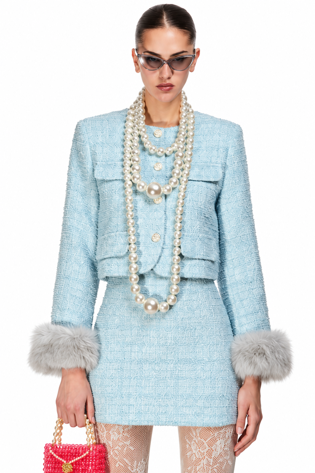 A model wearing a light blue tweed mini skirt suit with faux fur trim, accessorized with multiple layers of pearl necklaces and sunglasses, holding a pink beaded handbag against a white background.