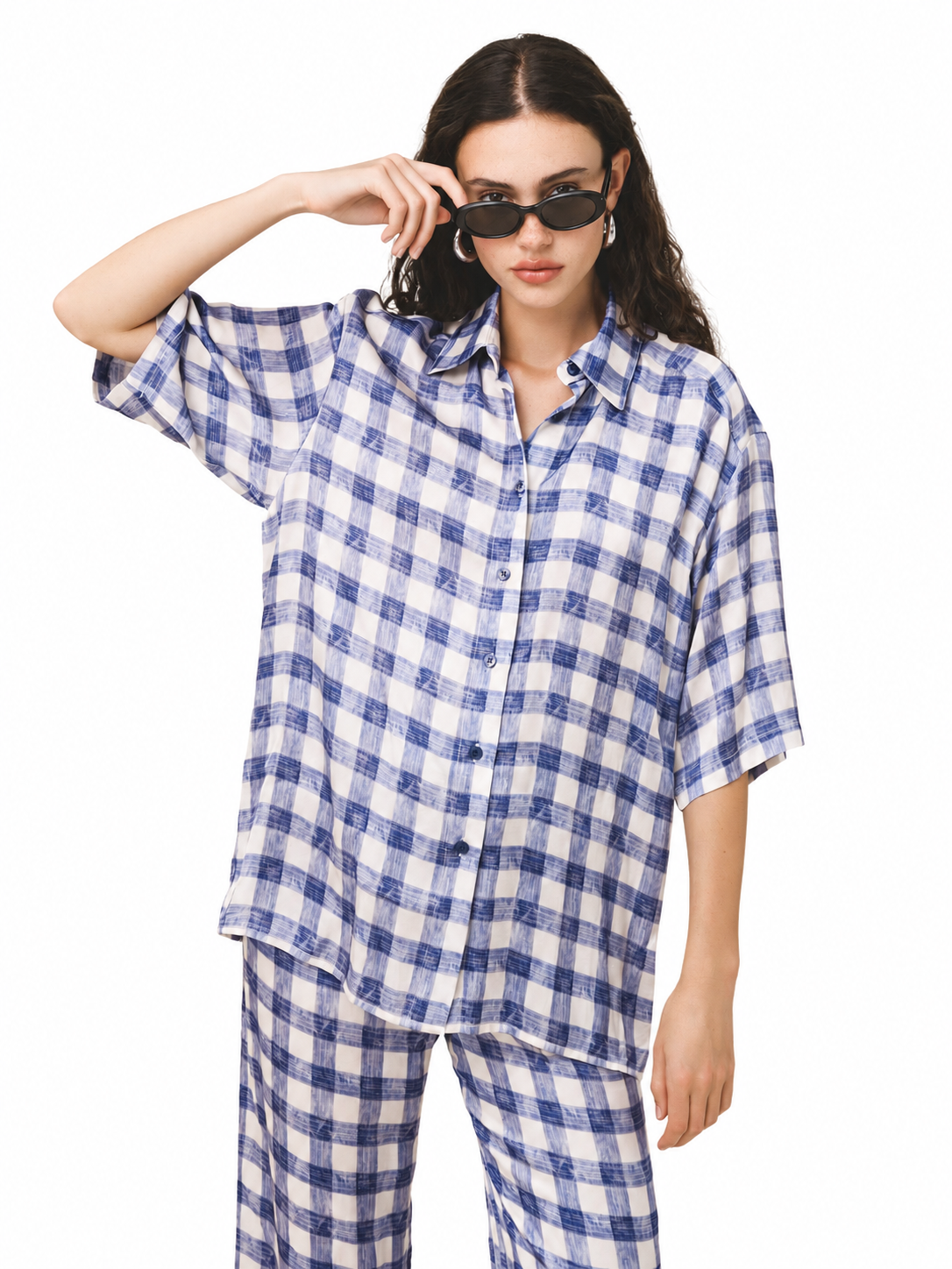 A woman modeling a matching blue and white gingham check pajama set, featuring an oversized short-sleeve button-up shirt and coordinating pants, against a plain white background.