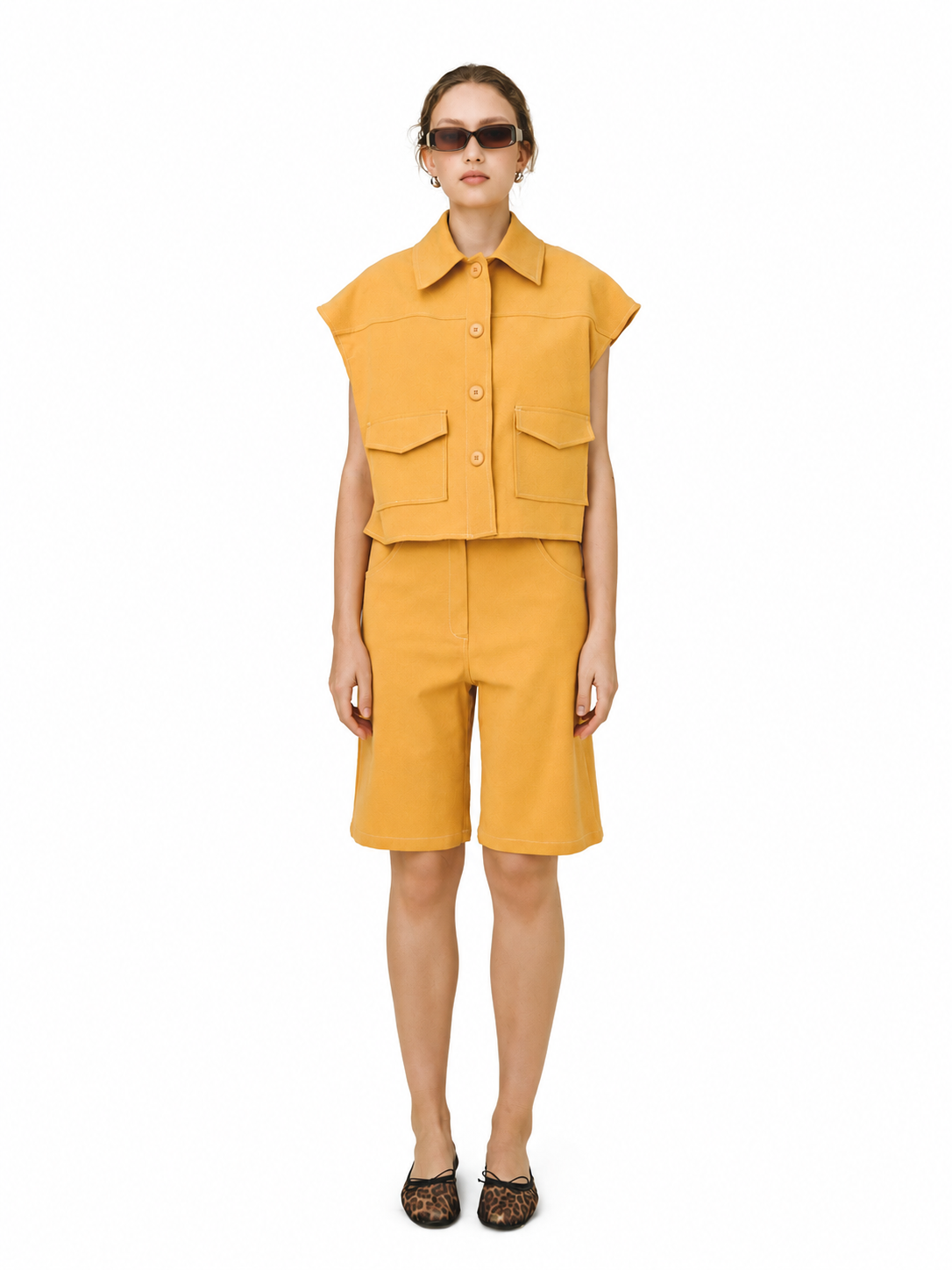 A model stands against a plain white background wearing a matching mustard yellow two-piece set consisting of a sleeveless collared button-front top with flap chest pockets and matching shorts. The model is also wearing dark sunglasses and leopard-print flat shoes.