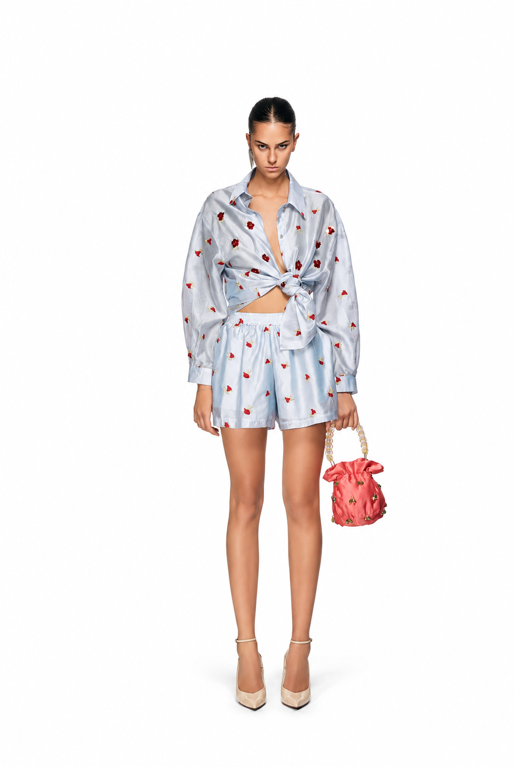 A model stands against a white studio background wearing a light blue satin shirt and matching shorts set with a red strawberry print, tied at the waist, and holding a small red strawberry handbag.