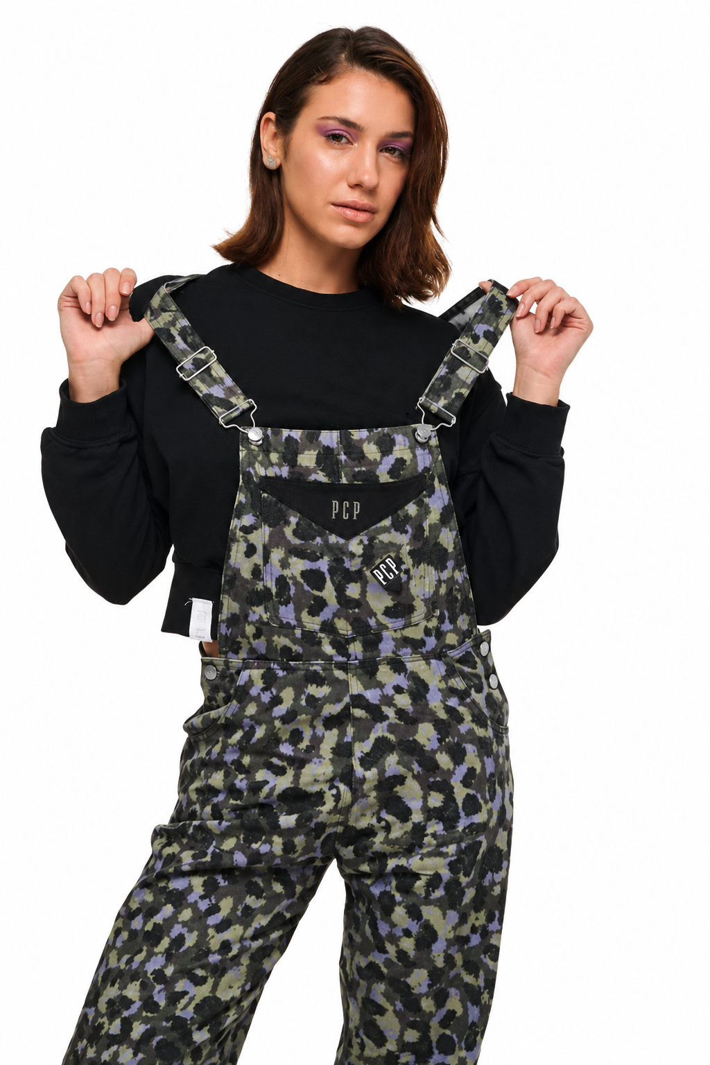 A model wearing green, black, and purple camouflage overall pants over a black long-sleeve shirt, holding the shoulder straps of the overalls against a white background.