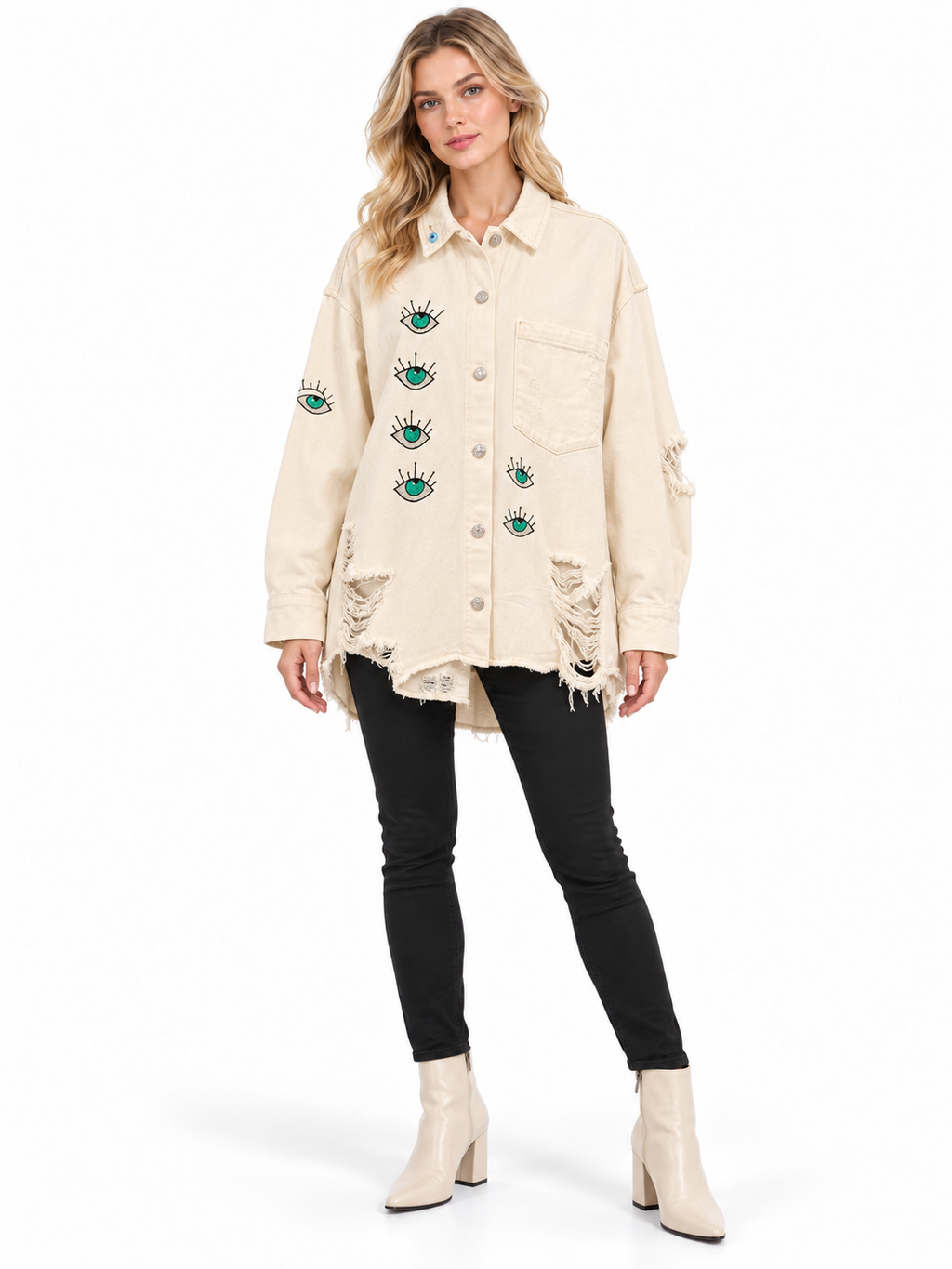 A woman modeling a distressed off-white denim jacket with embroidered green and blue eyes on the front and sleeves, paired with black skinny jeans and cream ankle boots, against a plain white background.