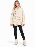 A woman modeling a distressed off-white denim jacket with embroidered green and blue eyes on the front and sleeves, paired with black skinny jeans and cream ankle boots, against a plain white background.