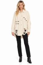 A woman modeling a distressed off-white button-up shirt jacket with ripped details and decorative patches, paired with black pants and black ankle boots, on a white studio background.
