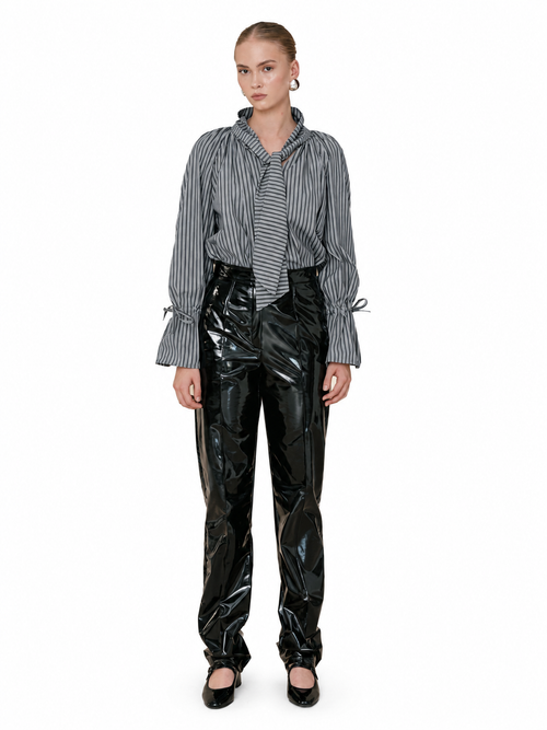 A model stands against a plain white background wearing a black and white vertically striped long-sleeve shirt with tie details at the cuffs, paired with shiny black patent leather pants and black shoes.