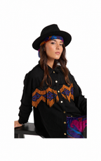 A woman wearing a black wide-brim hat, a colorful patterned headband, a black button-front jacket with orange and purple geometric fringe embroidery across the chest, and a purple patterned skirt, posed against a solid white background.