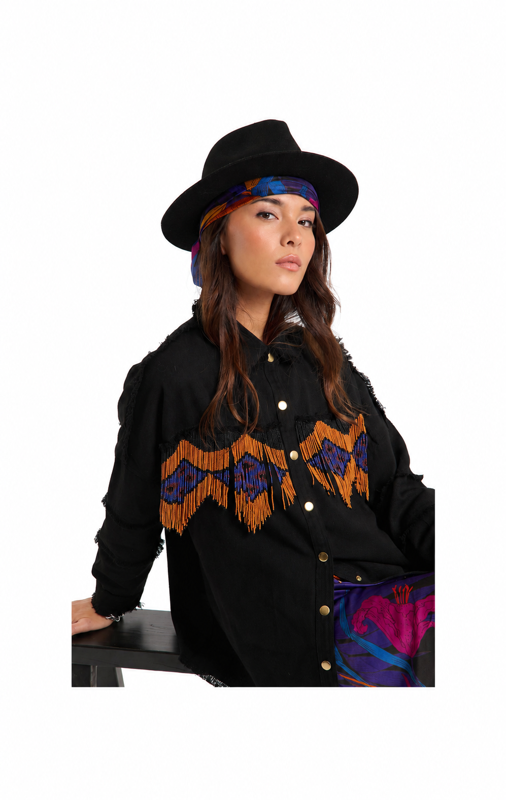 A woman wearing a black wide-brim hat, a colorful patterned headband, a black button-front jacket with orange and purple geometric fringe embroidery across the chest, and a purple patterned skirt, posed against a solid white background.