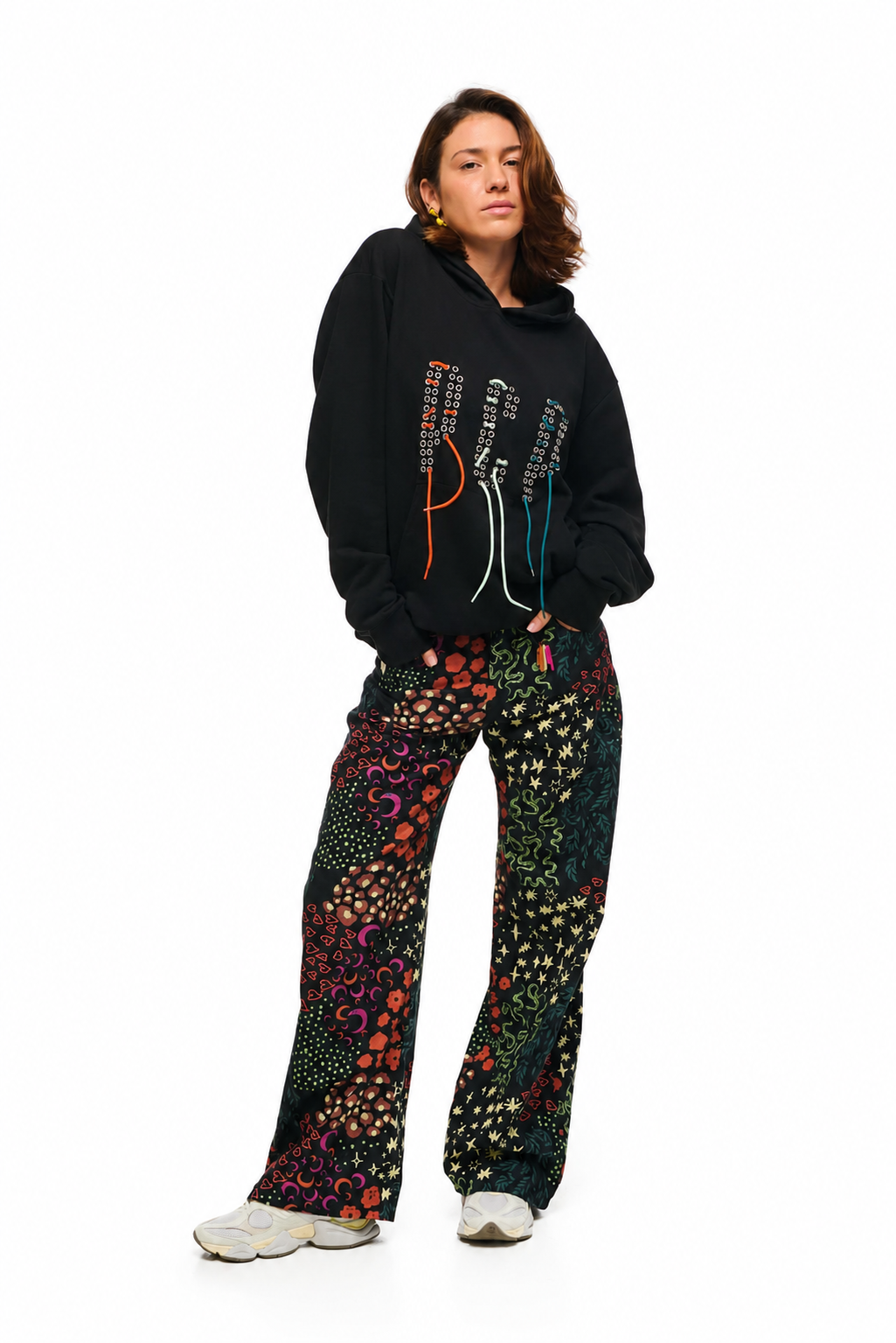 A woman modeling a black hoodie with colorful embroidered vertical lettering and matching patterned wide-leg pants, standing against a plain white background.