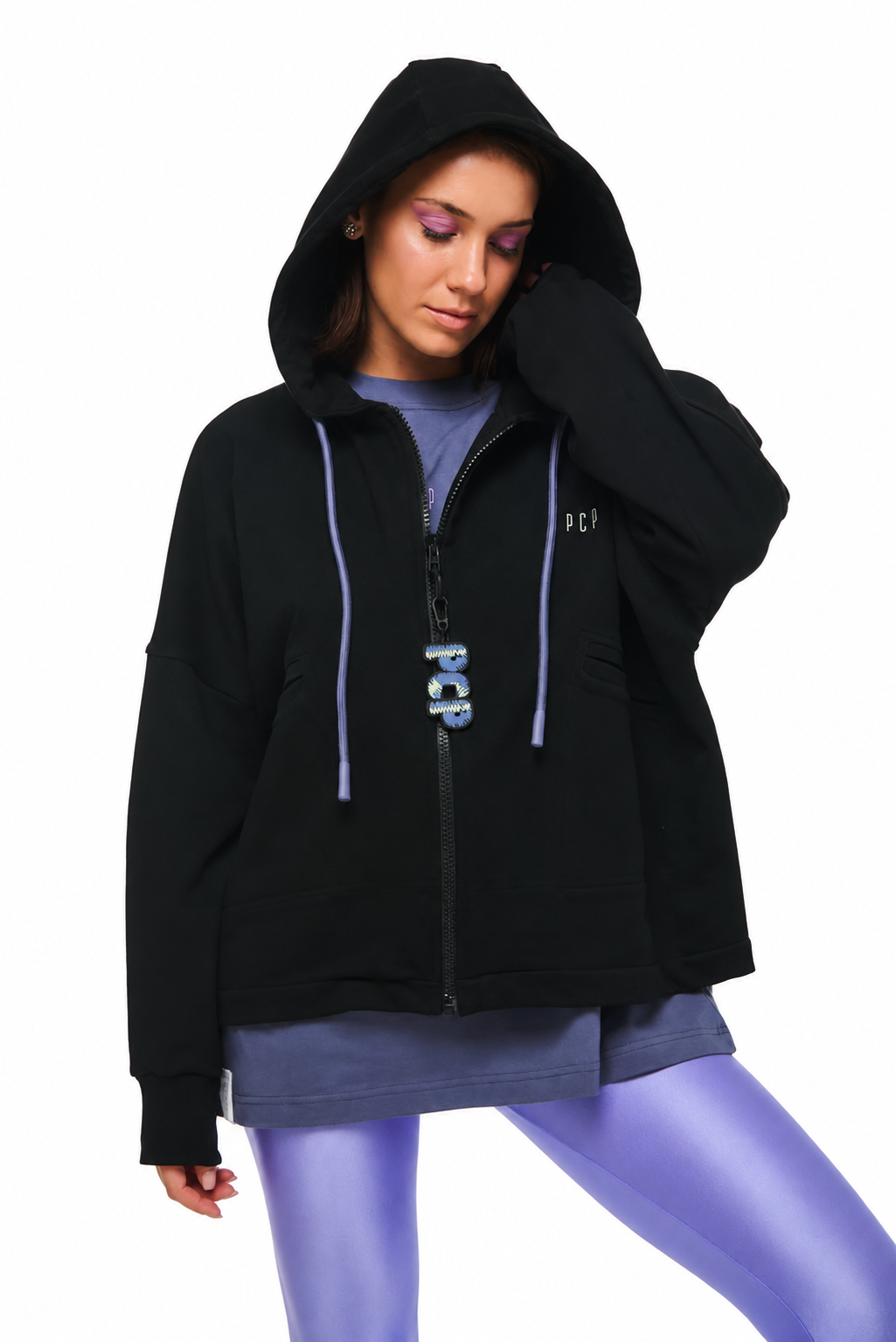 A woman modeling a black zip-up hoodie with light purple drawstrings and a matching purple 'PCP' logo on the front, worn over a purple top and purple leggings, against a plain white background.