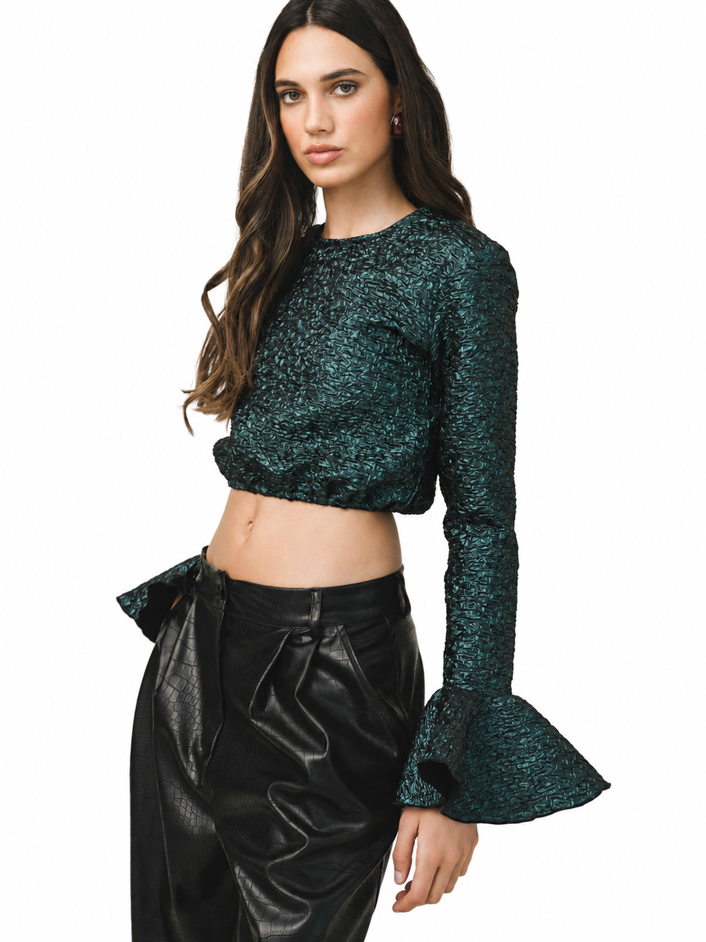 A woman modeling a dark green glittery long-sleeve crop top with flared cuffs, paired with black faux-leather pants, against a plain white background.