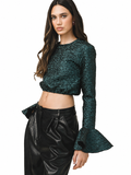 A woman modeling a dark green glittery long-sleeve crop top with flared cuffs, paired with black faux-leather pants, against a plain white background.
